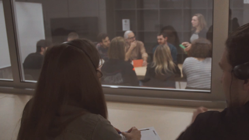 People participating in a focus group while two members of Food for Tomorrow are taking notes behind the spy glass.